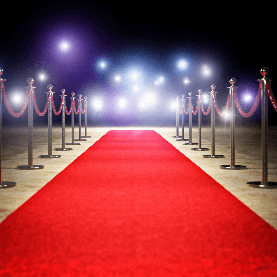 Red carpet lined with stanchions and ropes, illuminated by bright lights in a glamorous setting.